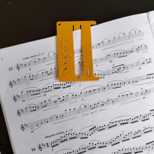 Peut inclure: Un serre-page en plastique jaune moutarde avec une découpe en forme de U et diverses perforations circulaires et rectangulaires. L'outil porte l'inscription 25mm sur sa surface et est posé sur une partition de musique en papier blanc.
