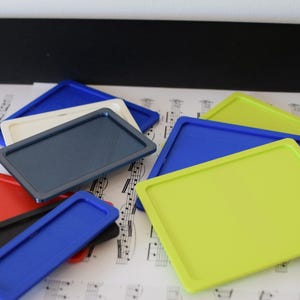 Peut inclure: Plusieurs plateaux rectangulaires colorés, bleus, verts, rouges, noirs et blancs. Les plateaux sont disposés sur une surface avec des notes de musique en arrière-plan. Les plateaux sont probablement destinés à organiser de petits objets.