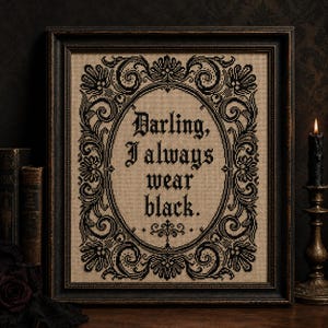 May include: A framed cross-stitch embroidery on tan fabric featuring a black ornate oval border and the text "Darling, I always wear black." The piece is enclosed in a dark, distressed wooden frame with a rectangular profile and decorative edges.