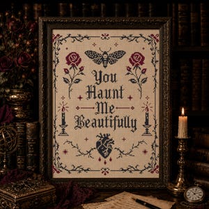 Può includere: Un ricamo a punto croce incorniciato con il testo You Haunt Me Beautifully in lettere nere. Il design include una falena, due rose rosse, due candele accese e un cuore anatomico, il tutto racchiuso in un bordo nero decorativo.