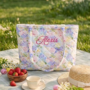Puede incluir: Un bolso tote acolchado con un estampado floral en tonos pastel de rosa, amarillo y azul. El nombre Alexis está bordado en cursiva rosa en la parte central frontal. El bolso tiene dos asas de tela a juego y una forma rectangular.