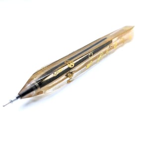 May include: A mechanical pencil with a clear, gold-toned body. The pencil features black stripes and gold-colored decorative gears and other small shapes. The pencil tip is silver and the lead is visible.