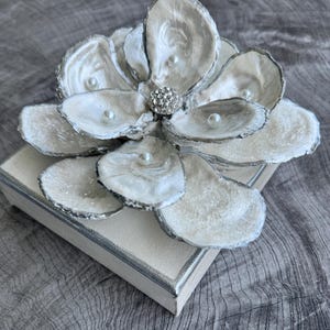 May include: A decorative flower made from oyster shells, painted white with silver edges, and adorned with faux pearls and a rhinestone center. The flower sits atop a rectangular box with a silver trim.