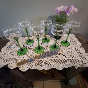 May include: Set of six clear wine glasses with green stems and bases. Each glass features a white floral design around the bowl. The glasses are arranged on a white lace doily, with a decorative fan and a small potted plant in the background.