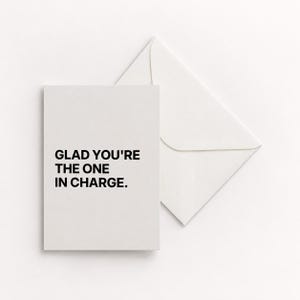 May include: A rectangular off-white greeting card featuring the black printed text GLAD YOU'RE THE ONE IN CHARGE. The card is positioned next to a matching off-white envelope with a triangular flap, both made of a smooth, matte paper material.