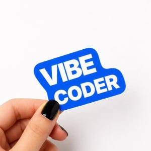 May include: A blue sticker with the words "VIBE CODER" in white. The sticker is held by a hand with a black painted fingernail against a white background.