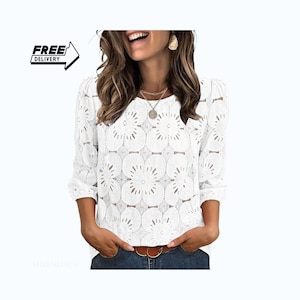 Puede incluir: Una blusa blanca de manga larga con un patrón de encaje floral en toda la prenda, bordes festoneados, cuello redondo y mangas abullonadas fruncidas. Se lleva con un cinturón marrón y vaqueros azules. El texto en la imagen dice FREE DELIVERY.