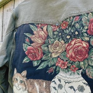 May include: An olive green jacket featuring a large rectangular tapestry panel sewn onto the back. The panel displays a dark blue background with a woven design of a brown tabby cat lying next to a white vase filled with red and yellow roses.