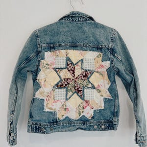 May include: A light blue acid-wash denim jacket featuring a large, square-shaped patchwork quilt panel sewn onto the back. The quilt displays a central star pattern composed of floral, yellow striped, and grey gingham fabric pieces with frayed edges.