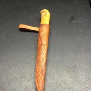 May include: A light brown leather knife sheath with a smooth, pale yellow wooden handle protruding from the top. The sheath features embossed geometric diamond patterns, a circular stamp with the letter M, and a leather belt loop attachment.