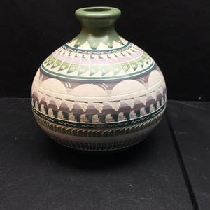 May include: A round ceramic vase with a narrow neck and a wide body. The surface features horizontal bands of geometric patterns in shades of sage green, lavender, white, and dark gray, with a matte finish and textured carved details.