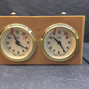 May include: A tan analog chess clock with two circular clock faces, gold-toned bezels, and black hands. Each face features the text USCF and Made in Germany. The device has two small metal buttons on the top for controlling the timing mechanism.
