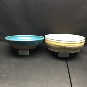 May include: Two ceramic bowls are displayed side by side. The bowl on the left features a vibrant turquoise blue glaze with a smooth finish. The bowl on the right has a white interior and a textured exterior with horizontal bands of yellow and brown.