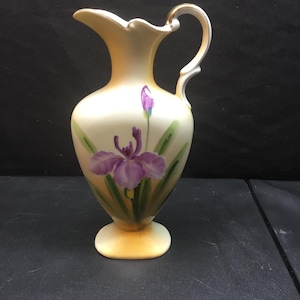 May include: A cream-colored porcelain pitcher featuring a hand-painted purple iris flower with green leaves. The pitcher has a curved handle with gold trim, a scalloped rim, and a soft yellow gradient finish at the base and neck, standing on a pedestal.