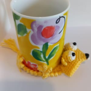 May include: A yellow ceramic mug with a floral design in purple, red, and green, resting on a yellow crocheted dog-shaped coaster. The mug has a white interior and a smooth, glossy finish. The coaster has black eyes and a black nose.