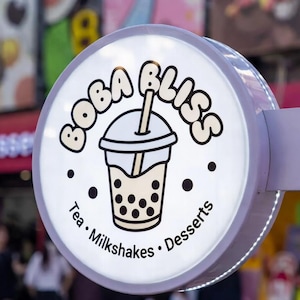 May include: A round, illuminated sign for "Boba Bliss" featuring a graphic of a boba tea drink. The sign also includes the words "Tea • Milkshakes • Desserts" in a circular design around the drink image.