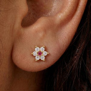 May include: A gold-colored flower-shaped stud earring. The earring features six white gemstones surrounding a central red gemstone. The earring is worn on an ear.