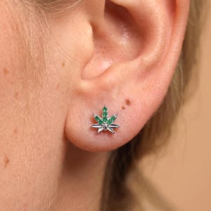 May include: A silver stud earring shaped like a cannabis leaf, featuring small, bright green stones embedded along the surface of the leaf design. The earring is positioned on the earlobe, showcasing a metallic finish and a detailed botanical shape.