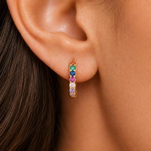 May include: A small gold hoop earring featuring a row of seven round, multi-colored gemstones. The stones are arranged in a vertical line, displaying shades of orange, green, blue, pink, white, light purple, and a final small pink accent.