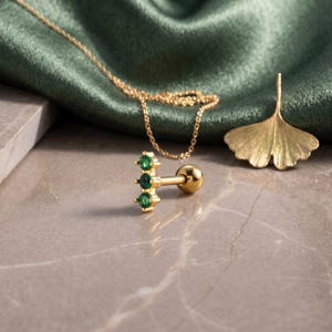 May include: A gold-tone barbell earring featuring three small, round, green stones set in a vertical row. The earring has a smooth, spherical backing. The piece is displayed alongside a gold-tone ginkgo leaf charm and a delicate gold-tone chain.