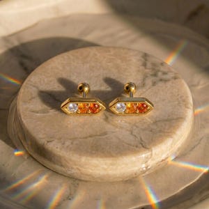 May include: A pair of gold-colored stud earrings. Each earring features a geometric design with a clear, orange, and orange-red gemstone arrangement. The earrings are set on a light-colored, circular surface.