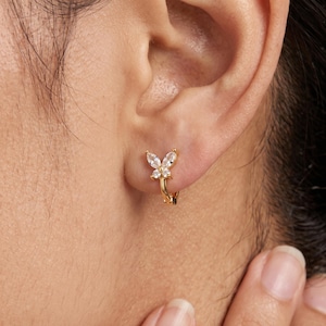 May include: A gold hoop earring featuring a butterfly-shaped charm adorned with clear, marquise-cut and round-cut stones. The earring is secured through the earlobe, showcasing a metallic gold finish and sparkling white gemstone accents.