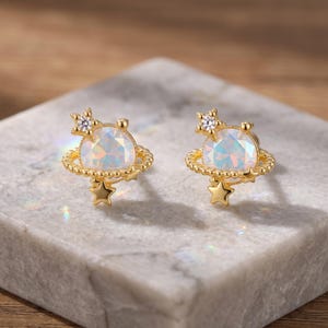 May include: A pair of gold-tone stud earrings shaped like planets with rings. Each earring features a central iridescent round stone, a small clear crystal star accent at the top, and a dangling gold star charm at the bottom of the ring.