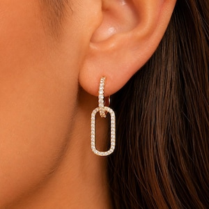 May include: Gold-toned hoop earrings with a rectangular pendant. The earrings are encrusted with small, clear gemstones, creating a sparkling effect. The design is modern and elegant, suitable for various occasions.