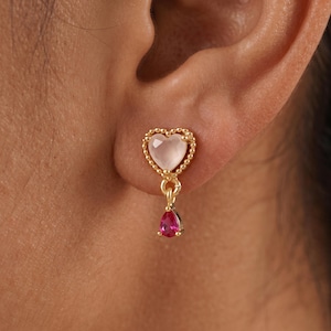 May include: A gold-toned stud earring featuring a heart-shaped pale pink stone framed by a beaded border. A small, teardrop-shaped vibrant pink stone hangs from the base of the heart, creating a delicate drop design that rests against the earlobe.