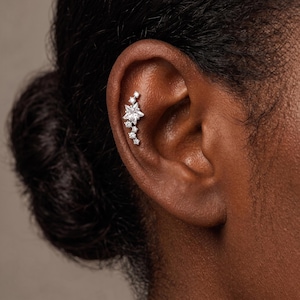 May include: A silver-toned cartilage earring featuring a cluster of clear, sparkling stones arranged in a vertical, star-inspired design. The jewelry piece consists of one central star-shaped stone surrounded by several smaller round stones.