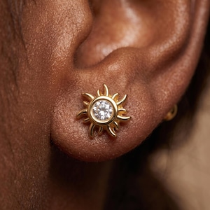 May include: A gold stud earring shaped like a sun with a central round clear gemstone. The metal frame features radiating curved points surrounding the stone, creating a sunburst design that sits flush against the earlobe for a bright appearance.