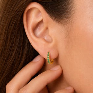 May include: A small gold hoop earring featuring a row of tiny, vibrant green gemstones set along the front curve. The earring is circular in shape with a polished metallic finish, providing a bright contrast against the embedded green stones.