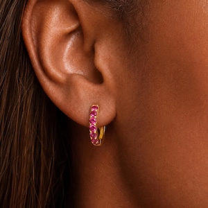 May include: A small gold hoop earring featuring a row of round, vibrant pink gemstones set along the front curve. The earring is secured through the earlobe, showcasing a polished metallic finish and a series of evenly spaced, bright pink stones.