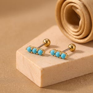 Könnte beinhalten: Ein Paar Barbell-Ohrringe mit drei kleinen, runden, leuchtend türkisfarbenen Perlen in einer horizontalen Reihe. Jeder Ohrring verfügt über einen glatten, polierten goldfarbenen Metallstift und einen Kugelverschluss für Piercings.