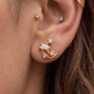 Peut inclure: Une boucle d'oreille à clou dorée au design incurvé inspiré d'une constellation. Le bijou est orné de trois petites pierres rondes transparentes et d'une pierre centrale taille baguette, serties dans une fine monture en métal doré perlée.