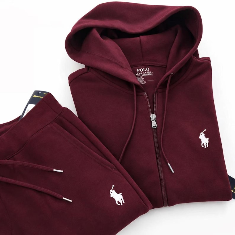 May include: A burgundy Polo Ralph Lauren tracksuit set. The set includes a zip-up hoodie with a drawstring hood and matching sweatpants. Both pieces feature the Polo Ralph Lauren logo in white. The hoodie has a full zipper closure.