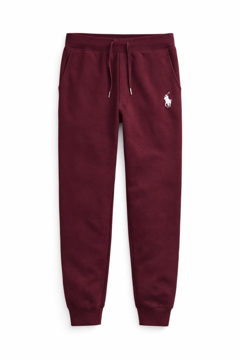 May include: Burgundy-colored sweatpants with a drawstring waist and side pockets. A white polo player logo is embroidered on the left side. The pants have a relaxed fit and cuffed ankles, suitable for casual wear.