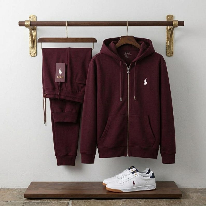 May include: A burgundy zip-up hoodie and matching sweatpants set with a white logo. White sneakers with navy blue accents and a brown sole are displayed below the clothing. The set is hanging on a wooden rack.