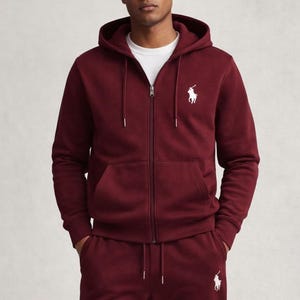 May include: Burgundy zip-up hoodie and matching sweatpants set. The hoodie features a drawstring hood, a front zipper, and a white logo on the chest. The sweatpants also have a white logo. The outfit is made of a soft material.