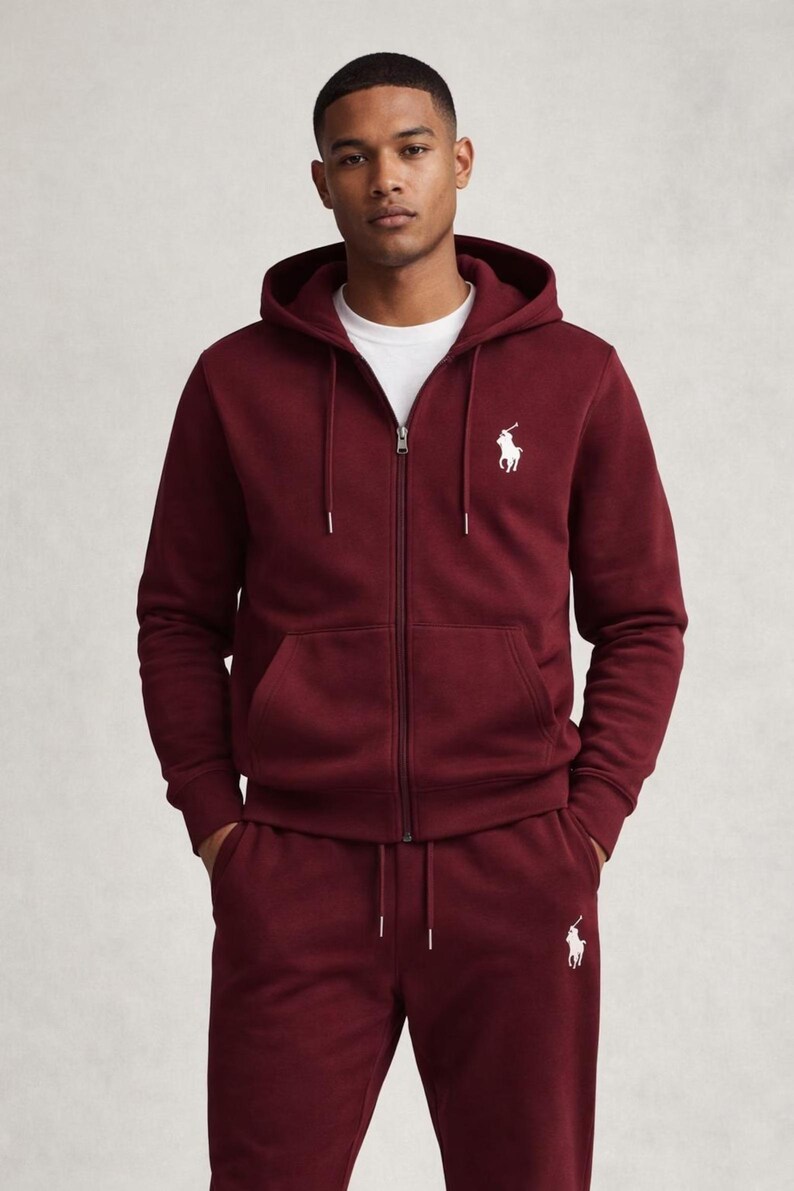 May include: Burgundy zip-up hoodie and matching sweatpants set. The hoodie features a drawstring hood, a front zipper, and a white logo on the chest. The sweatpants also have a white logo. The outfit is made of a soft material.