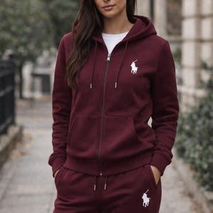 May include: Burgundy zip-up hoodie and matching sweatpants. The hoodie features a white logo on the chest and a drawstring hood. The sweatpants also have the white logo on the left leg. The outfit is made of a soft material.