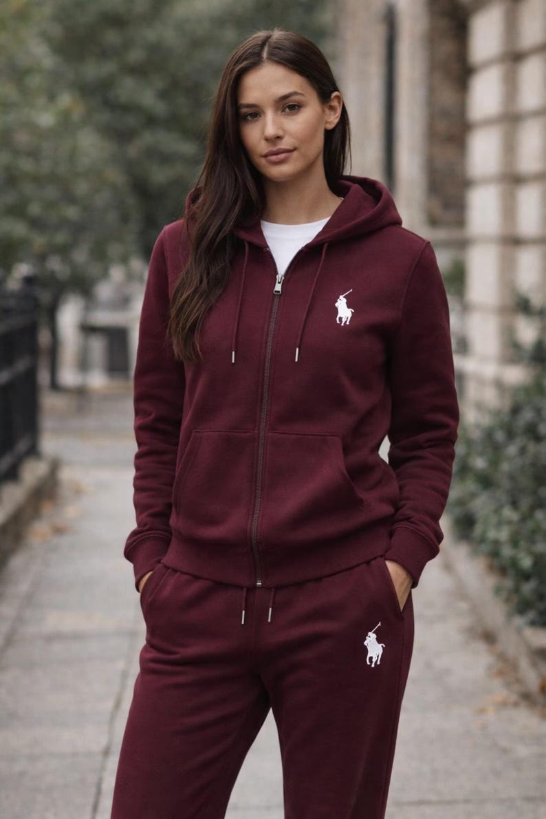 May include: Burgundy zip-up hoodie and matching sweatpants. The hoodie features a white logo on the chest and a drawstring hood. The sweatpants also have the white logo on the left leg. The outfit is made of a soft material.