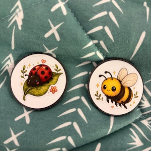 May include: Two round, decorative pins featuring cartoon insects. One pin shows a red ladybug on a green leaf, and the other shows a yellow and black bee with wings. The pins are on a teal fabric.