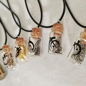 Könnte beinhalten: Fünf Glasfläschchen-Halsketten an schwarzen Kordeln. Jede durchsichtige Flasche ist mit einem Korkstopfen verschlossen und enthält metallische Elemente wie Goldflocken, Silberketten oder Draht. Auf der Vorderseite jedes Glasfläschchens ist ein schwarzes Mondsichelsymbol aufgedruckt.