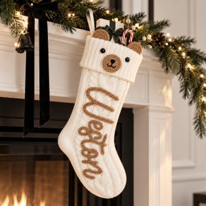 Puede incluir: Un calcetín navideño de punto trenzado color crema con un diseño de cara de oso en el puño y orejas de ganchillo marrones. El nombre Weston está bordado con hilo marrón en el centro. El calcetín contiene un pequeño regalo y un bastón de caramelo.