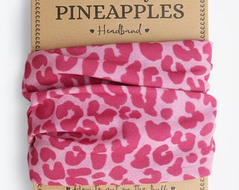Puede incluir: Una diadema con estampado de leopardo rosa y fucsia. La diadema se muestra en un respaldo de cartón con el texto "The Curly Pineapples Headband". El embalaje también incluye instrucciones sobre cómo usar la diadema.