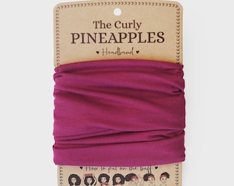 Puede incluir: Una diadema magenta oscuro de The Curly Pineapples, exhibida en un respaldo de cartón. El texto en el empaque dice "The Curly Pineapples Headband". La diadema es de color sólido y parece estar hecha de una tela suave y elástica.