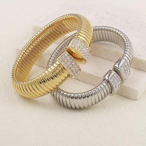May include: Two metal bracelets, one gold-toned and one silver-toned, are displayed. Both feature a ribbed design and are adorned with a rectangular clasp embellished with small, clear crystals. The bracelets are positioned on a light-colored surface.