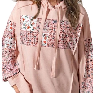 May include: A light pink hoodie with floral patchwork details. The sleeves and front panel feature a mix of floral patterns in shades of pink, red, and white. The hoodie has a drawstring and a kangaroo pocket.