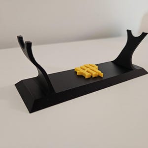 Peut inclure: Un présentoir noir avec deux supports incurvés et une base rectangulaire. Un petit emblème jaune est placé sur la surface du présentoir. Le présentoir est conçu pour contenir un objet, probablement une épée ou un autre objet de collection.